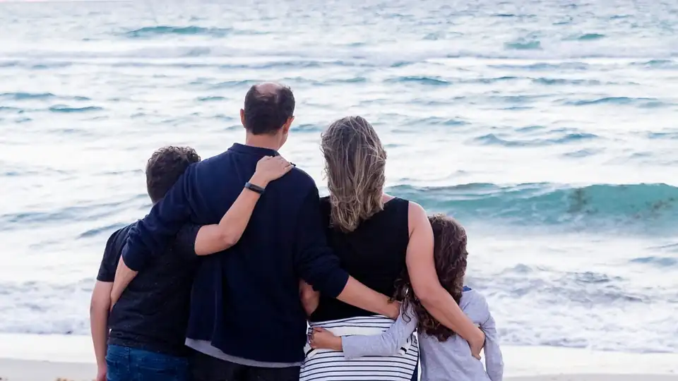 Family of four standing together on a beach, facing the ocean with their arms around one another